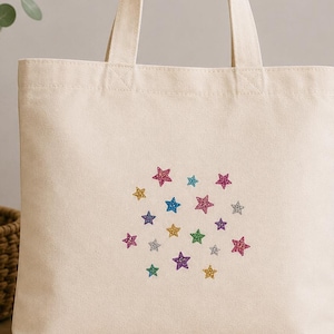 May include: A cream-coloured canvas tote bag featuring a central cluster of small, multi-coloured glittery star icons. The stars are arranged in a scattered pattern and appear in various shades including pink, blue, yellow, purple, green, and silver.
