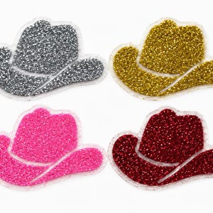 May include: A set of four glittery cowboy hat patches arranged in a grid. The patches are silver, gold, bright pink, and deep red. Each hat-shaped applique features a clear, raised border and a dense, sparkling texture across the entire surface.