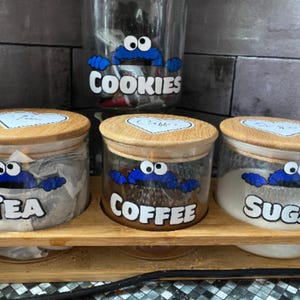 May include: Three glass jars with bamboo lids and cookie monster themed labels. The labels read "Tea", "Coffee", and "Sugar".
