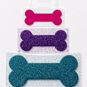 May include: Three bone-shaped acrylic tags arranged vertically. The top tag is solid hot pink. The middle tag is purple glitter on a clear rectangular base. The bottom tag is teal glitter on a clear rectangular base with a curved edge.
