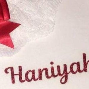 May include: White fabric featuring a red satin bow and the name "Haniyah" in red glitter lettering. The fabric has a delicate lace trim. The design is simple and elegant.