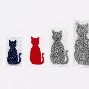 May include: Five cat-shaped iron-on patches arranged in a row. The set includes a small white glitter cat, a dark blue glitter cat, a solid red cat, a medium silver glitter cat, and a large silver glitter cat, all on rectangular backings.
