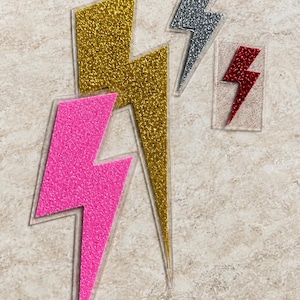 May include: Four lightning bolt patches with glittery finishes. One large gold bolt, one large hot pink bolt, one medium silver bolt, and one small red bolt. Each bolt is mounted on a clear, transparent backing that follows the shape of the bolt.