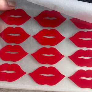 May include: A sheet of red lip-shaped stickers arranged in a grid pattern on a white backing paper. Each sticker features a bold, solid red colour with a cutout line representing the mouth opening, creating a repeating graphic design of lips.