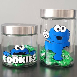 May include: Two glass cookie jars with silver lids. The smaller jar has a Cookie Monster graphic with the word "COOKIES" underneath. The larger jar has a Cookie Monster graphic holding a cookie.