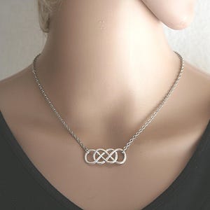 Può includere: Una collana color argento con una serie di simboli dell'infinito intrecciati. Il ciondolo è su una catena delicata, creando un design minimalista ed elegante. La collana è esposta su un manichino.
