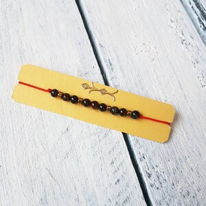 Può includere: Un braccialetto di corda rossa con perline verde scuro e rosse. Il braccialetto è su una carta gialla con un design dorato.