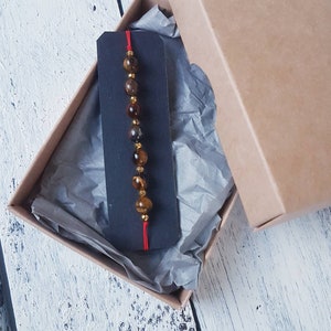 Può includere: Un braccialetto con perline marroni e dorate con un filo rosso legato alle estremità, esposto su una carta nera all'interno di una scatola marrone con carta velina.