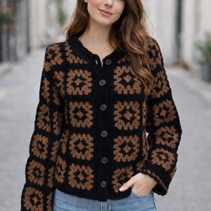 Può includere: Un cardigan all'uncinetto nero e marrone con chiusura a bottoni. Il cardigan presenta un motivo quadrato marrone e nero, con maniche lunghe e scollatura rotonda. I bottoni sono marroni. La persona indossa jeans blu.