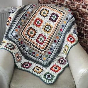 Può includere: Una coperta all'uncinetto colorata con un motivo a quadrati della nonna in tonalità di rosso, arancione, giallo, verde, blu e bianco. La coperta è drappeggiata su una poltrona bianca.