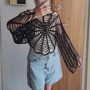 May include: A black crochet top featuring an intricate spider web pattern that radiates from the centre. The garment has long, loose sleeves and an open-knit construction, worn over a light-coloured base layer with light blue denim shorts.