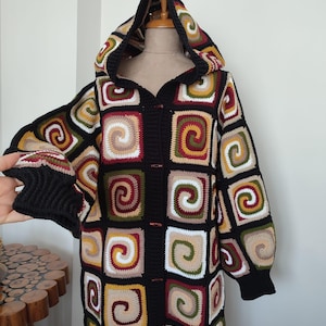 Puede incluir: Cárdigan largo con capucha tejido a ganchillo, con base negra y patrones cuadrados coloridos. Los cuadrados presentan un diseño en espiral en tonos crema, marrón, verde y rojo. Las mangas y la capucha están ribeteadas en negro, con botones negros.