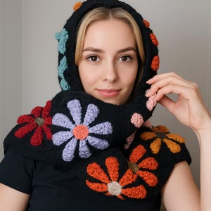 Puede incluir: Gorro de ganchillo negro con detalles de margaritas coloridas. El gorro presenta flores rojas, naranjas, moradas y celeste, con pompones naranjas en el borde superior. Se usa sobre una camiseta negra.
