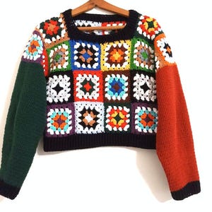 Può includere: Un maglione all'uncinetto colorato con sfondo nero e quadrati multicolori. Le maniche sono verdi e arancioni.