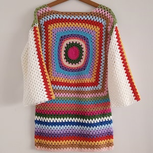 Puede incluir: Un top de crochet colorido con un patrón de rayas arcoíris y un gran diseño circular en el centro. El top tiene mangas largas con un ribete de crochet blanco.