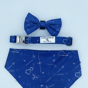 May include: A blue dog bandana with a white cloud and star pattern. The bandana has the text "CLOUD DOODLES EST 2021" on it. A matching blue bow tie and collar are also included.