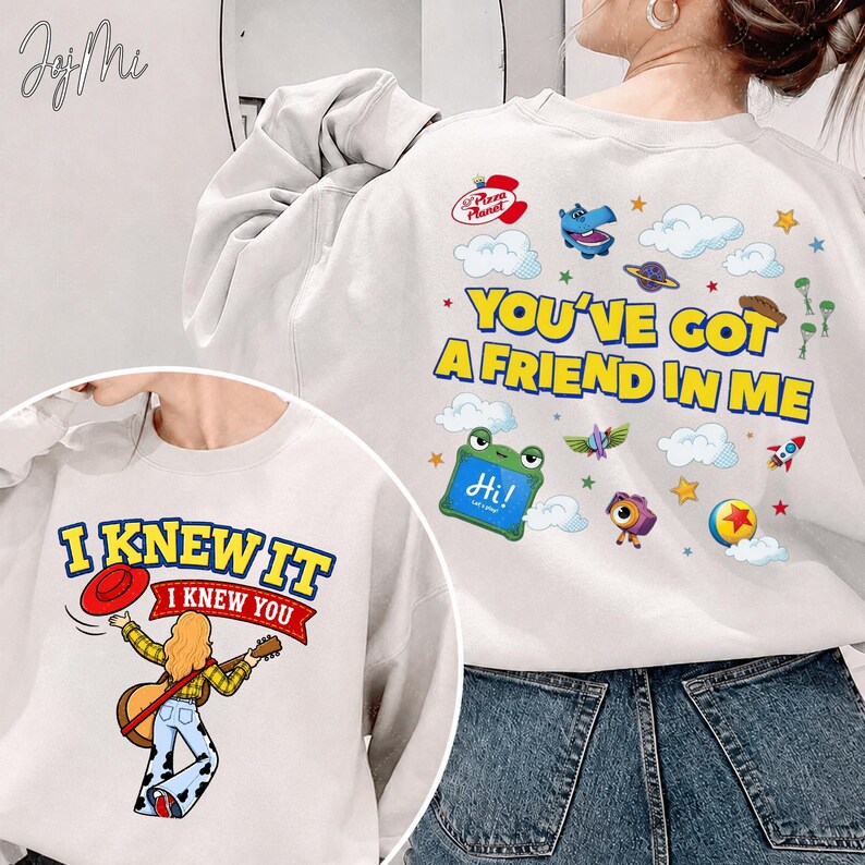 May include: A white crewneck sweatshirt featuring two graphic designs. The back displays the text "YOU'VE GOT A FRIEND IN ME" with colorful cartoon icons including clouds, a rocket, and a hippo. The front shows a person playing guitar with the text "I KNEW IT".