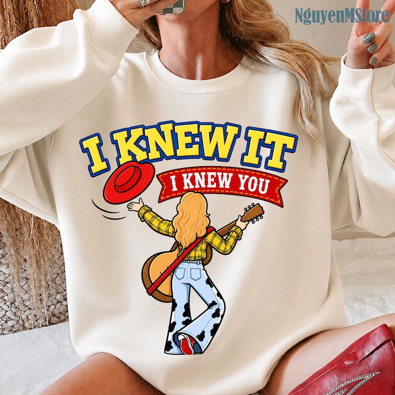 Peut inclure: Un sweat-shirt &agrave; col rond couleur cr&egrave;me avec un motif repr&eacute;sentant une personne aux longs cheveux blonds portant une chemise &agrave; carreaux jaune et un jean pattes d'eph &agrave; motif vache, tenant une guitare et un chapeau rouge, avec le texte I KNEW IT I KNEW YOU.