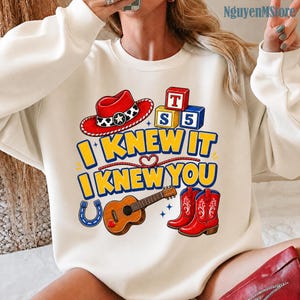 Peut inclure: Un sweat-shirt &agrave; col rond couleur cr&egrave;me avec une impression graphique repr&eacute;sentant un chapeau de cowboy rouge, des blocs alphabet, une guitare, un fer &agrave; cheval et des bottes de cowboy rouges. Le texte indique I KNEW IT I KNEW YOU en bleu et jaune.