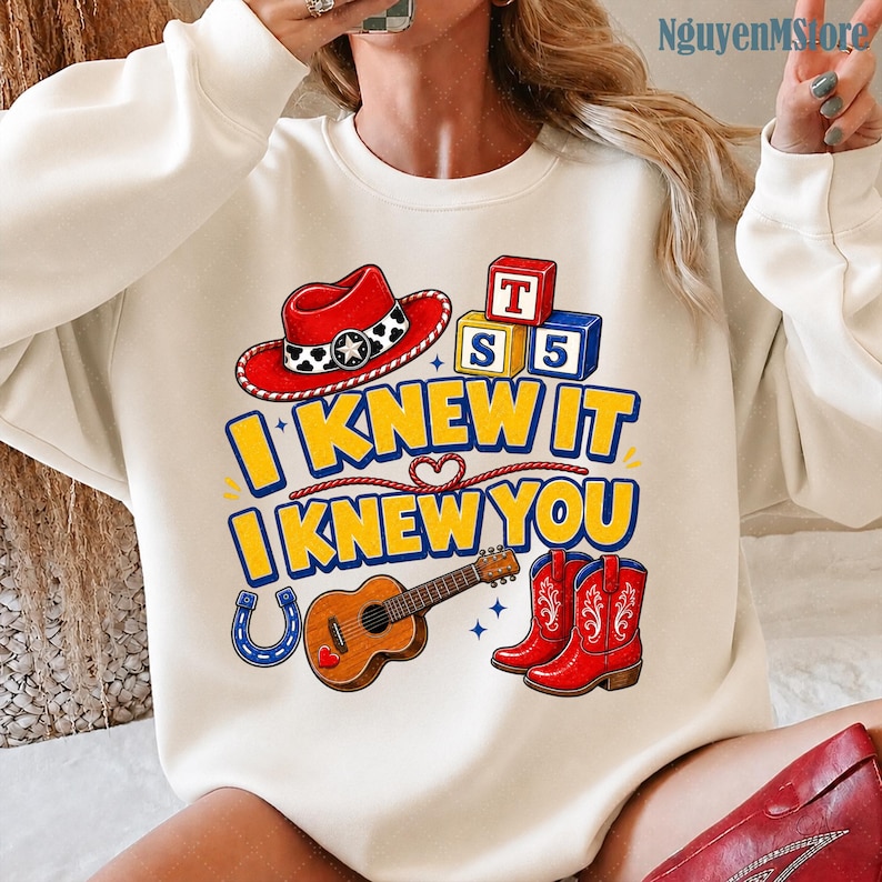 Peut inclure: Un sweat-shirt &agrave; col rond couleur cr&egrave;me avec une impression graphique repr&eacute;sentant un chapeau de cowboy rouge, des blocs alphabet, une guitare, un fer &agrave; cheval et des bottes de cowboy rouges. Le texte indique I KNEW IT I KNEW YOU en bleu et jaune.