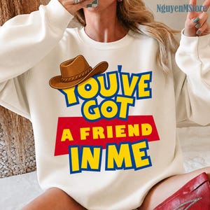 Peut inclure: Un sweat-shirt &agrave; col rond de couleur cr&egrave;me arborant un motif de chapeau de cowboy marron au-dessus du texte jaune et bleu "YOU'VE GOT A FRIEND IN ME" sur un fond rectangulaire rouge. Le motif est imprim&eacute; sur le devant du v&ecirc;tement &agrave; manches longues.