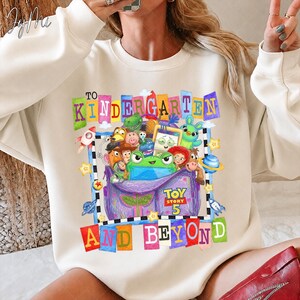 May include: A cream-colored crewneck sweatshirt featuring a colorful graphic print of various animated characters emerging from a purple backpack. The text reads TO KINDERGARTEN AND BEYOND with a TOY STORY 5 logo. The design includes stars and planets.