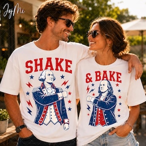 May include: Two white t-shirts featuring red and blue graphic prints. The left shirt displays George Washington with the text SHAKE, while the right shirt shows Benjamin Franklin with the text & BAKE. Both designs include red lightning bolts and blue stars.