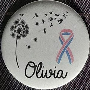 Peut inclure: Un bouton blanc avec un pissenlit noir, un ruban rose et bleu, et le nom Olivia en écriture cursive noire.
