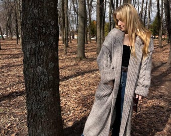 May include: A long, light gray cable-knit cardigan with pockets. The open-front sweater is worn over a black top and blue jeans. The person is wearing white sneakers and standing in a forest with bare trees.