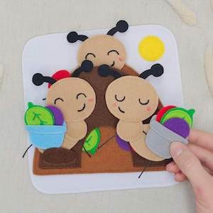 May include: A felt play mat featuring three smiling caterpillar figures with black antennae and carrying colourful felt food. The background includes a yellow sun and a brown hill, all on a white square.