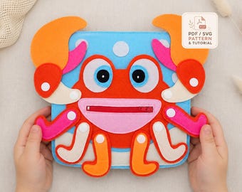 Pode incluir: Um projeto de artesanato em feltro em forma de caranguejo com corpo azul, pinças laranja e uma boca rosa com um fecho vermelho. O design inclui bolinhas brancas, olhos grandes e vários membros fixados com botões brancos.