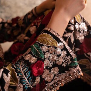 May include: Close-up of a black garment with intricate floral embroidery. The sleeves are adorned with colorful threadwork, including red roses, green leaves, and silver embellishments. The design features a detailed, ornate pattern.