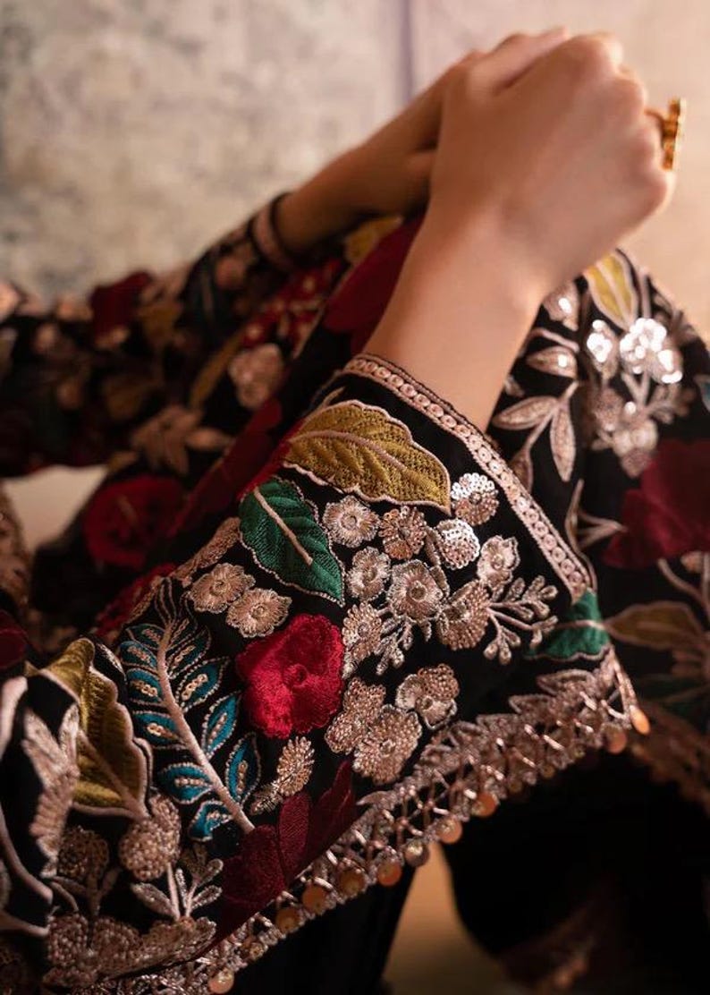 May include: Close-up of a black garment with intricate floral embroidery. The sleeves are adorned with colorful threadwork, including red roses, green leaves, and silver embellishments. The design features a detailed, ornate pattern.
