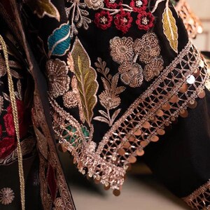 May include: Close-up of a black garment with intricate embroidery. The design features gold, red, and teal floral patterns, with gold sequins along the edges. The fabric appears to be a luxurious material.