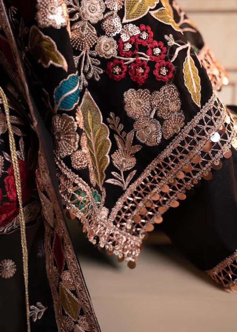 May include: Close-up of a black garment with intricate embroidery. The design features gold, red, and teal floral patterns, with gold sequins along the edges. The fabric appears to be a luxurious material.