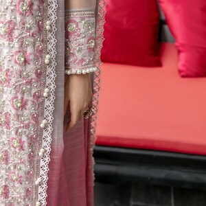 May include: A long, pink and gray dress with intricate embroidery and pearl accents. The dress has a lace trim and a sheer overlay. The dress is worn by a person standing in front of a red couch.