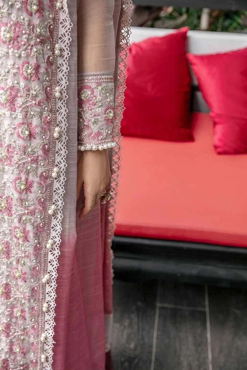 May include: A long, pink and gray dress with intricate embroidery and pearl accents. The dress has a lace trim and a sheer overlay. The dress is worn by a person standing in front of a red couch.