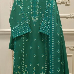 May include: Emerald green tunic and matching scarf set. The tunic features intricate white floral embroidery along the neckline, sleeves, and hem. The scarf is draped over the tunic, showcasing a coordinated design. The fabric appears to be lightweight.