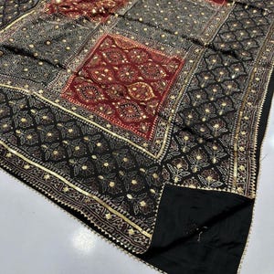 K&ouml;nnte beinhalten: Ein schwarz-rotes Textil mit Muster und goldenen Akzenten. Der Stoff hat ein Patchwork-Design mit floralen und geometrischen Mustern. Goldener Besatz umrandet die Kanten des Textils.