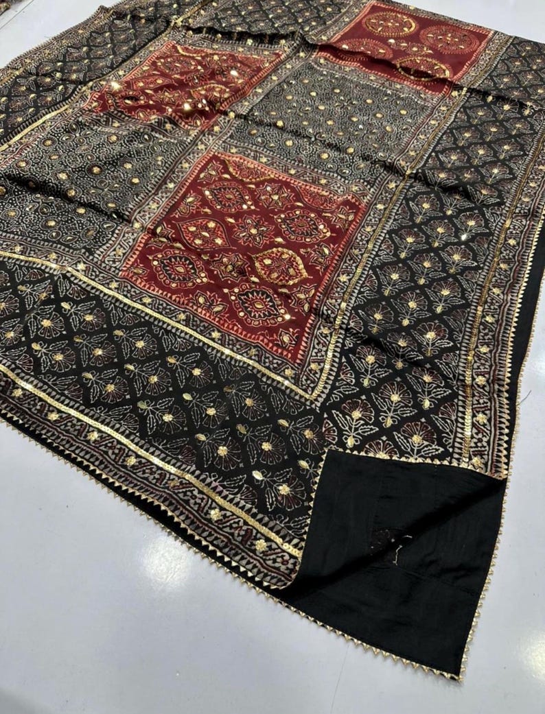 K&ouml;nnte beinhalten: Ein schwarz-rotes Textil mit Muster und goldenen Akzenten. Der Stoff hat ein Patchwork-Design mit floralen und geometrischen Mustern. Goldener Besatz umrandet die Kanten des Textils.