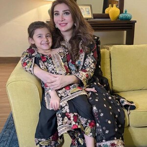 May include: A woman and a young girl are seated on a yellow sofa, both wearing black outfits with intricate floral embroidery in red, green, and gold. The woman's outfit includes a long tunic and pants, while the girl wears a similar ensemble. The background features a mirror and decorative items.
