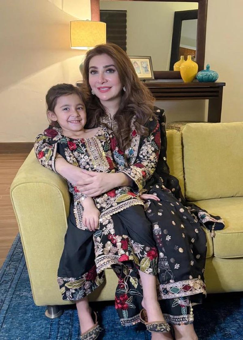 May include: A woman and a young girl are seated on a yellow sofa, both wearing black outfits with intricate floral embroidery in red, green, and gold. The woman's outfit includes a long tunic and pants, while the girl wears a similar ensemble. The background features a mirror and decorative items.