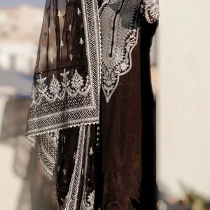 May include: A woman wearing a black and silver embroidered outfit with a sheer black dupatta. The outfit features intricate floral and geometric patterns. The woman is standing in front of a white wall.