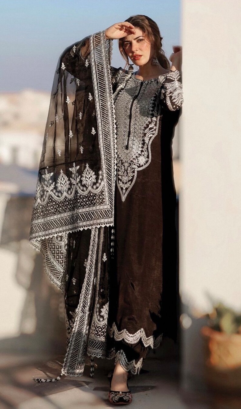 May include: A woman wearing a black and silver embroidered outfit with a sheer black dupatta. The outfit features intricate floral and geometric patterns. The woman is standing in front of a white wall.