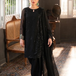 May include: Black embroidered formal wear ensemble. The outfit includes a long-sleeved tunic with intricate detailing, matching trousers, and a flowing scarf. The model is wearing green earrings and a red flower in their hair.
