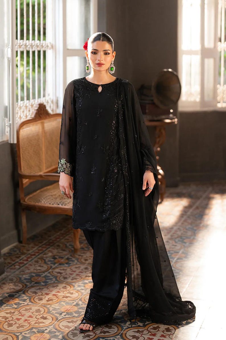 May include: Black embroidered formal wear ensemble. The outfit includes a long-sleeved tunic with intricate detailing, matching trousers, and a flowing scarf. The model is wearing green earrings and a red flower in their hair.