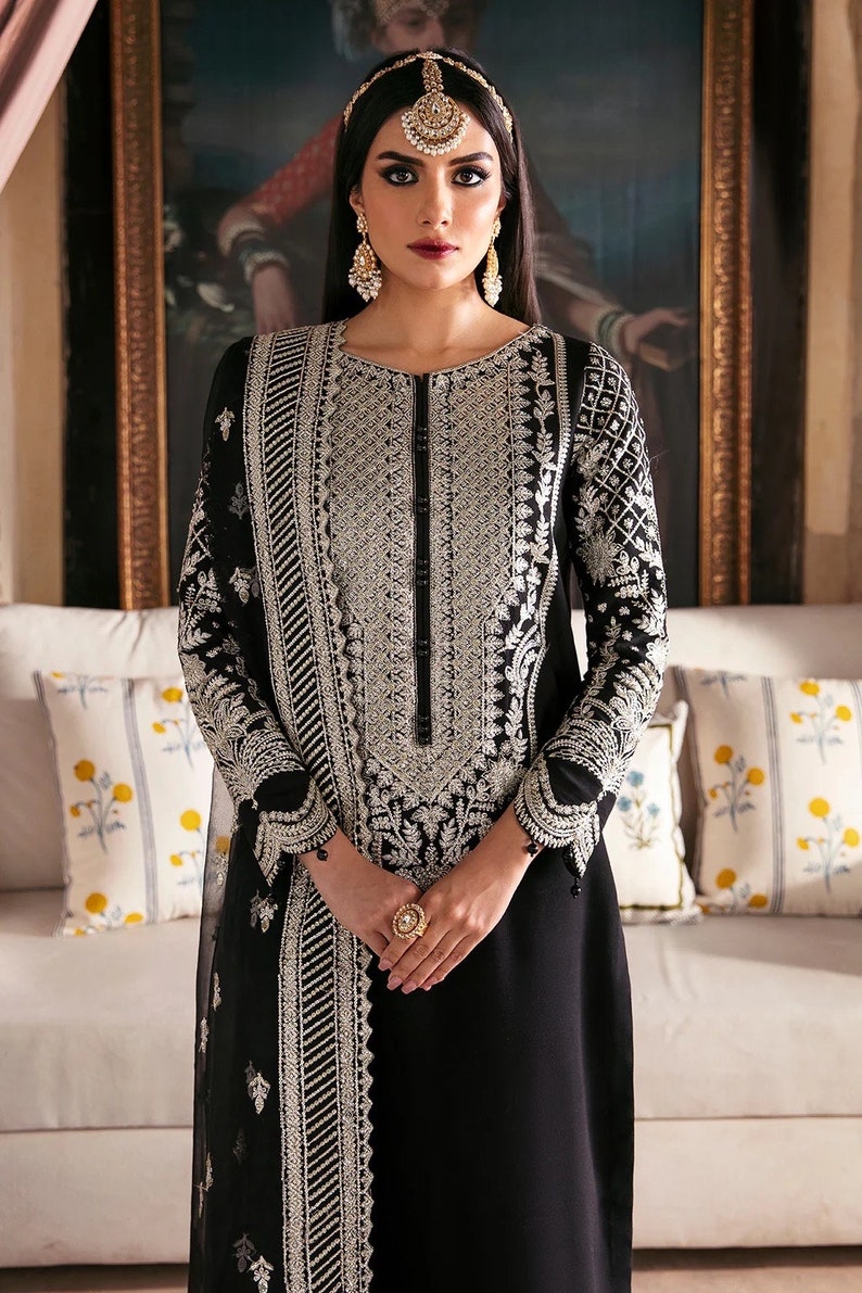 May include: A black long-sleeved dress with silver embroidery and a matching dupatta. The dress has a round neckline and a button closure. The model is wearing a gold headpiece and earrings.