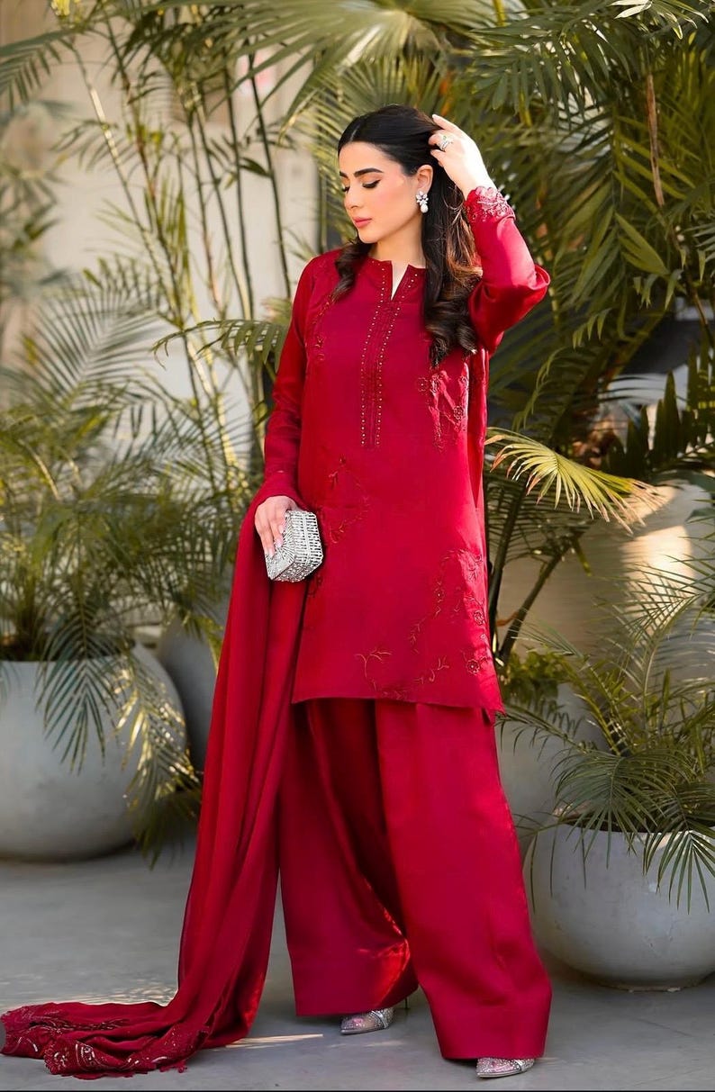 May include: A woman wearing a red embroidered dress with a matching dupatta. The dress has a long-sleeved top and wide-legged pants. She is holding a silver clutch.