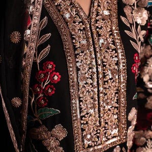 May include: Close-up of a black garment with intricate gold embroidery and sequin detailing. The design features floral patterns in red, gold, and green. The neckline is embellished with gold sequins.