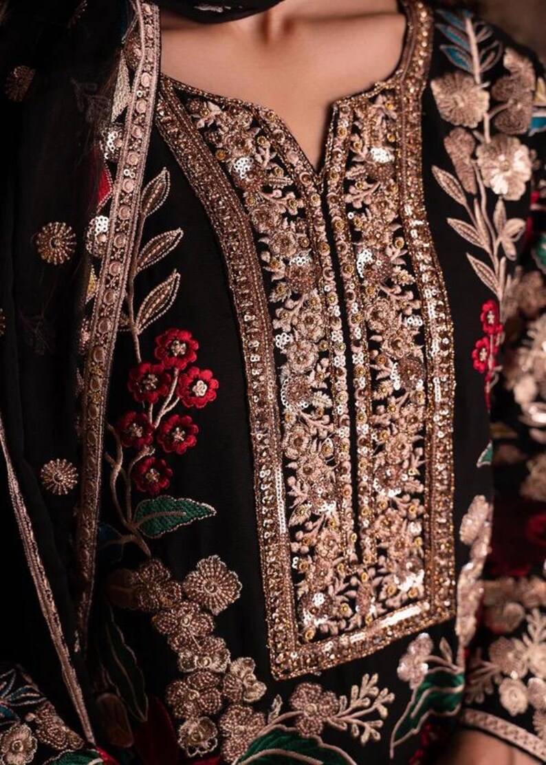 May include: Close-up of a black garment with intricate gold embroidery and sequin detailing. The design features floral patterns in red, gold, and green. The neckline is embellished with gold sequins.